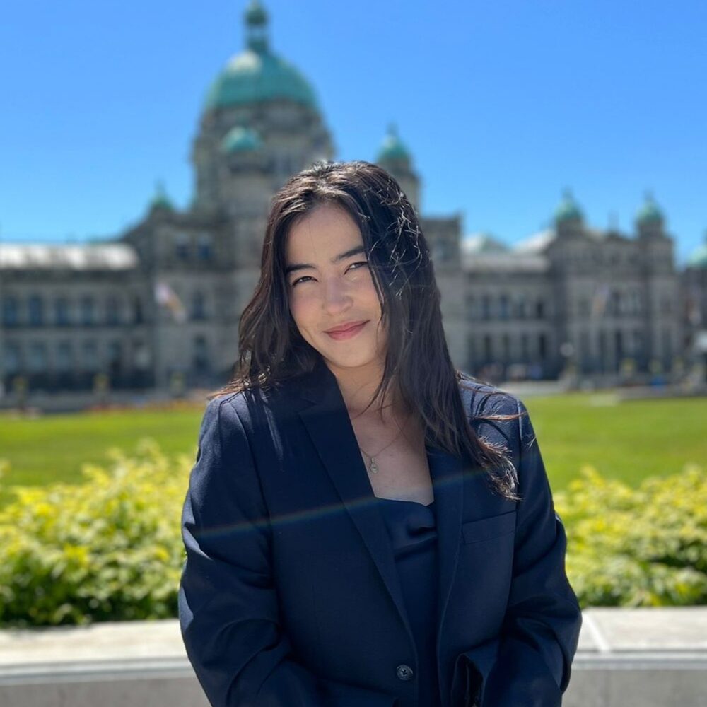 A headshot of Emily Burlie, Speech-Language Pathologist at Granville Island Speech. She stands in front of a green field with a large building in the background.