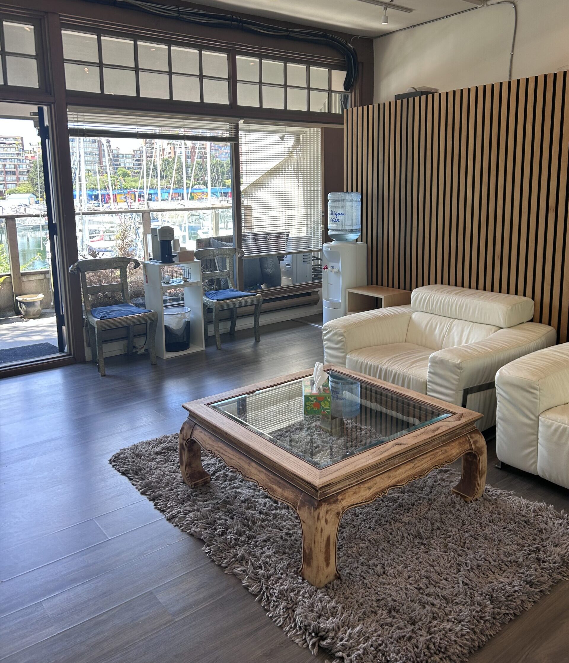 A photo of the waiting room of Granville Island Speech. The boats and pier of Granville Island are visible through the window. There are chairs, sofas, a coffee table, a water cooler, and a shelf in the waiting room.