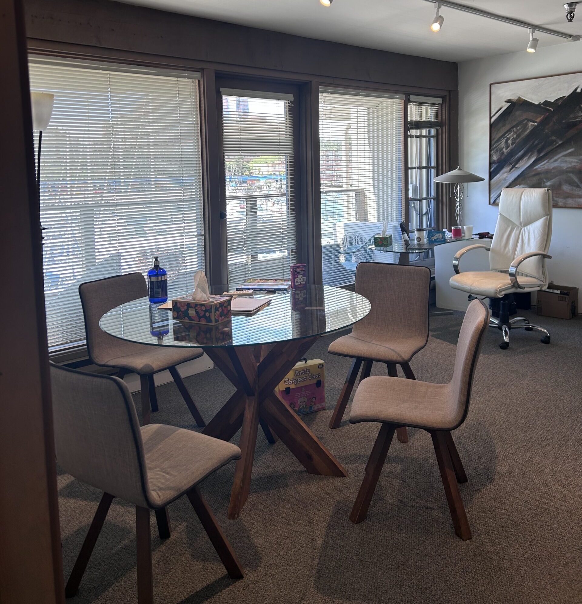 A photo of one of the rooms inside the Granville Island Speech clinic. There is a table in the middle of the room with four chairs around it. There are large floor to ceiling windows with the blinds partially closed. The floor is carpeted and there is a painting on the wall in the back. 