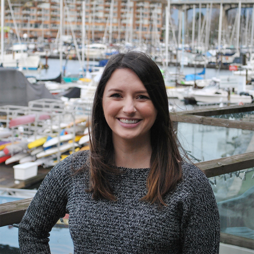 A headshot photo of Lauren Maynard, Speech-Language Pathologist at Granville Island Speech. She stands in front of boats and the pier at Granville Island outside of the clinic.