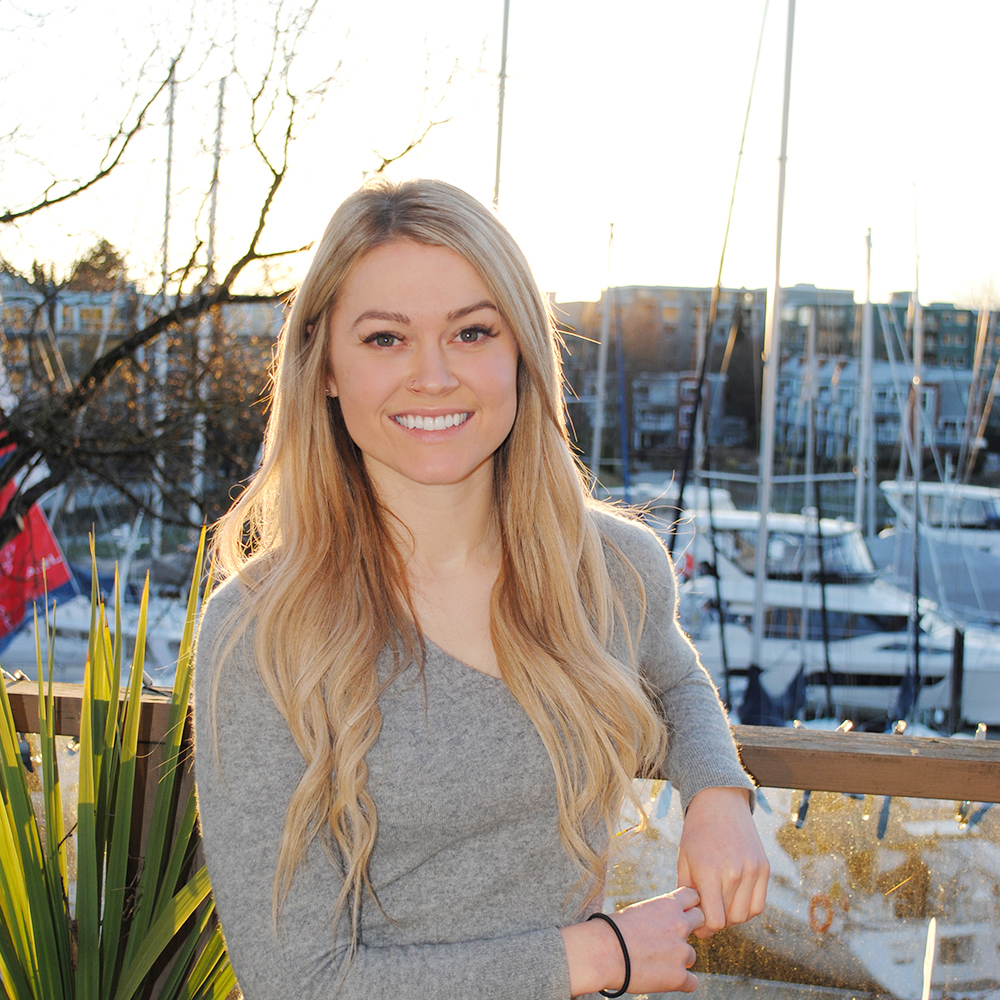 A headshot photo of Tara Nay, a Speech-Language Pathologist at Granville Island Speech. She stands outside of the clinic with boats and the pier in the background.