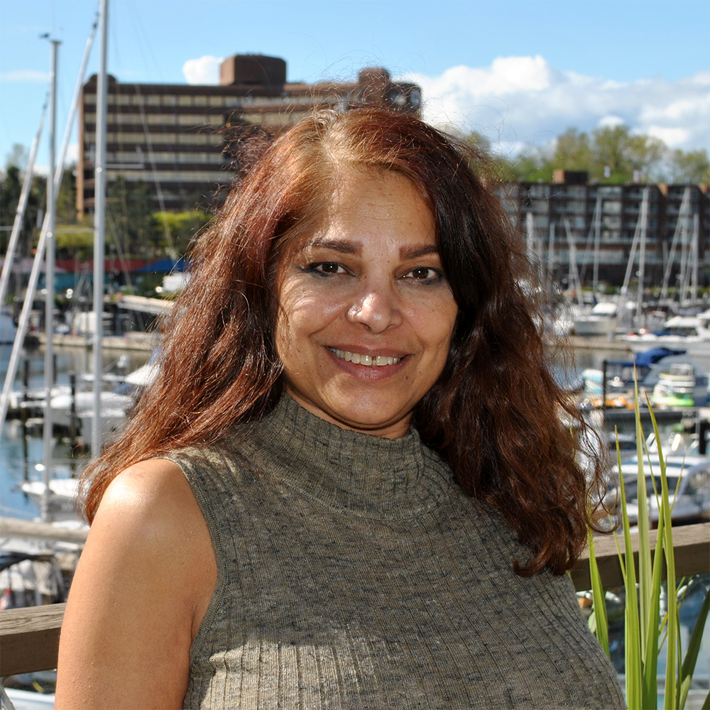 Headshot photo of Vinni Panikkar, Founder and Director of Granville Island Speech. She is standing in front of the clinic at Granville Island with boats and the pier in the background.