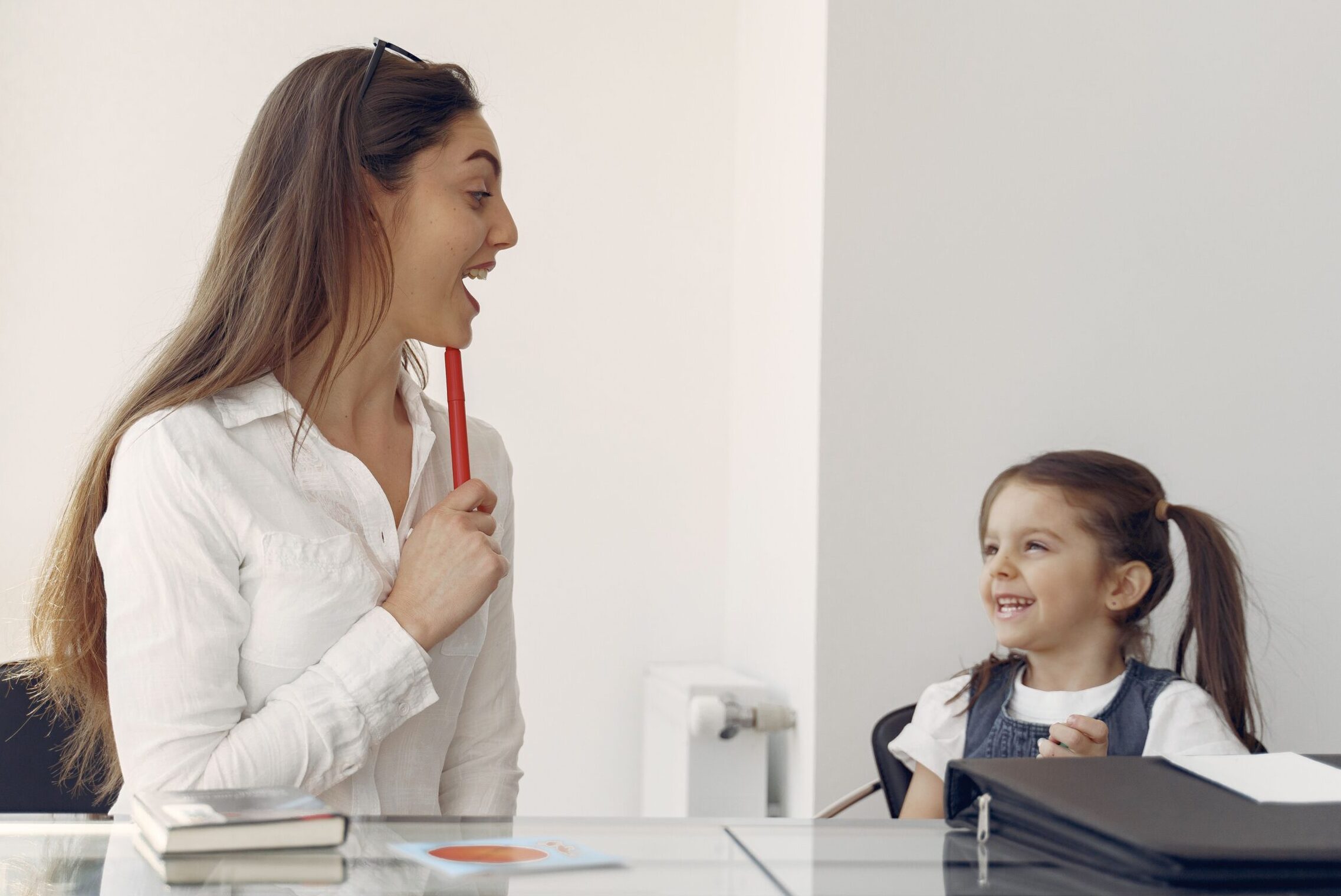 A speech therapist is sitting next to a young girl at a table, teaching her how to pronounce words.